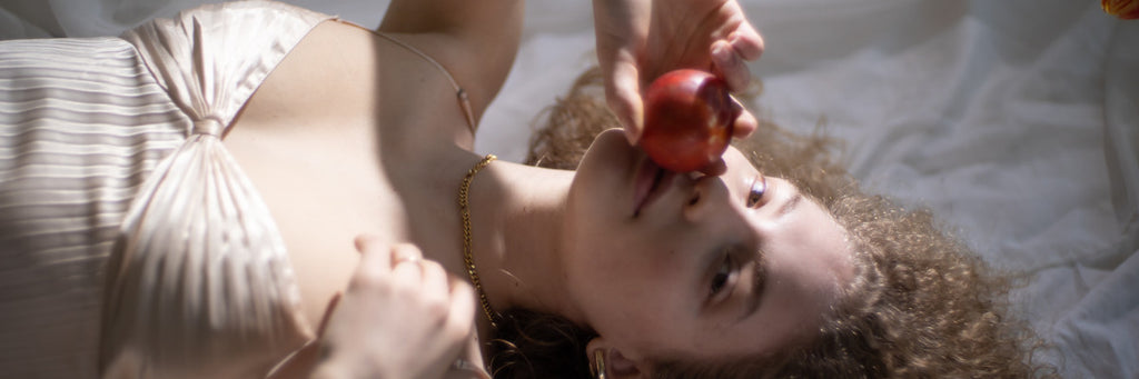Frau in elegantem Trägertop liegt auf dem Rücken, schaut in die Kamera und führt einen roten Apfel zu ihren Lippen.