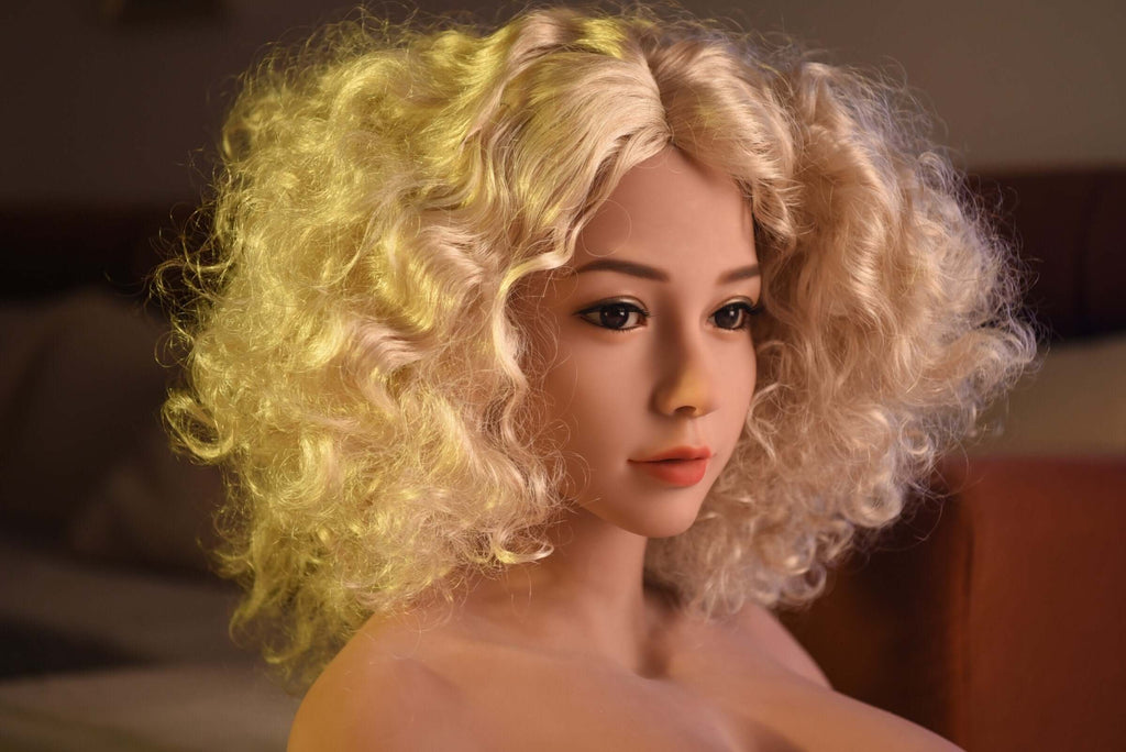 Blonder Lockenkopf Realdoll Sophia mit runden, großen, weichen Brüsten räkelt sich nackt in Schlafzimmer