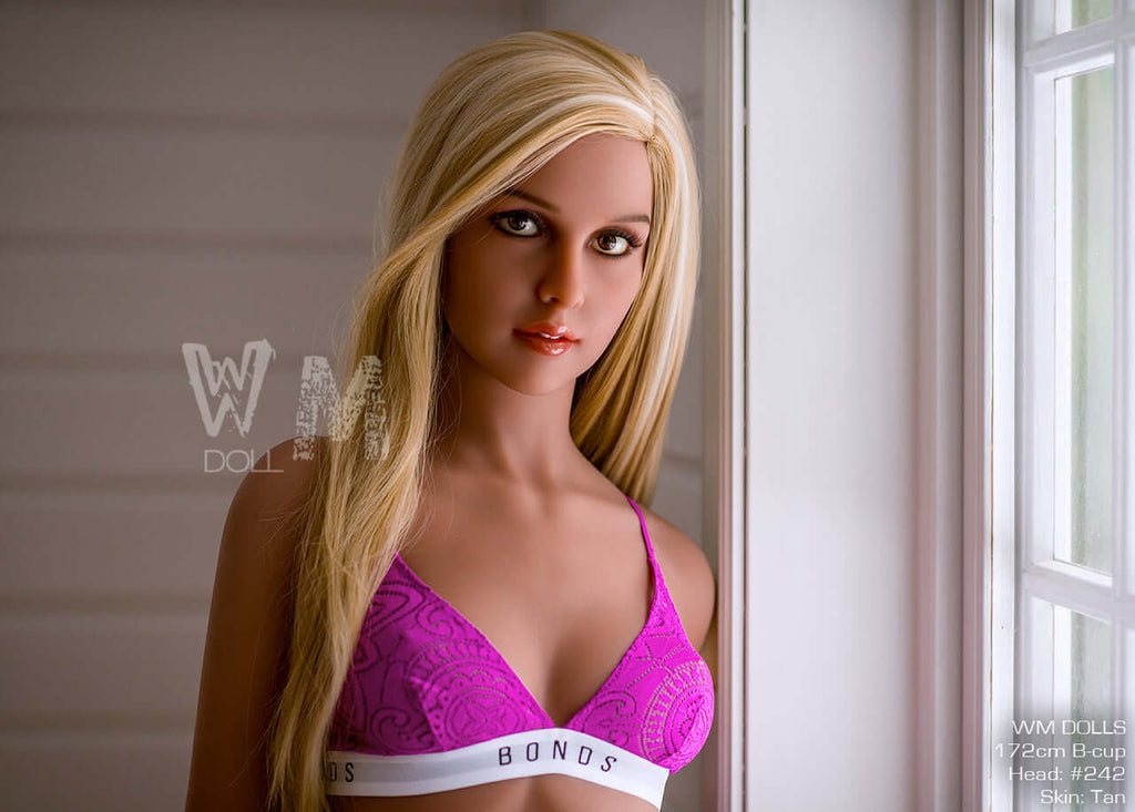 Sportliche, blonde Realdoll Juliette, mit runden Brüsten und kleinen rosa Nippeln, vor hellem Fenster